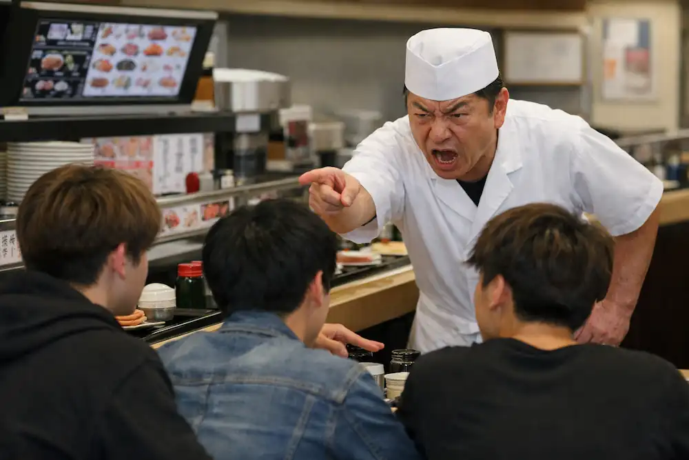 壽司店內，店員嚴肅地對年輕顧客表達不滿，呈現緊張的對話場面